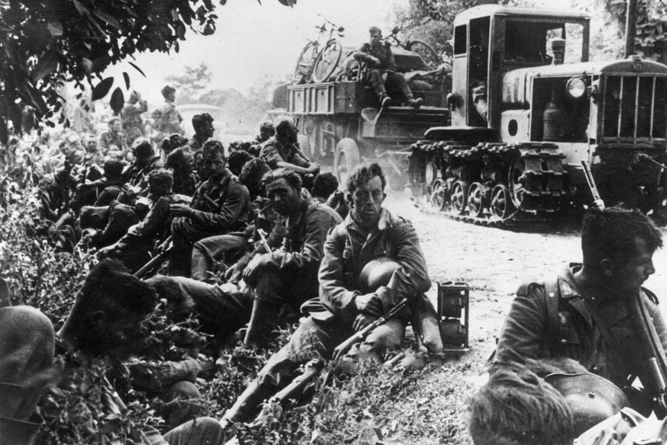 German soldiers rest on the side of the road somewhere in the USSR. A captured Soviet tractor SHTZ-NATI with a trailer is driving along the road