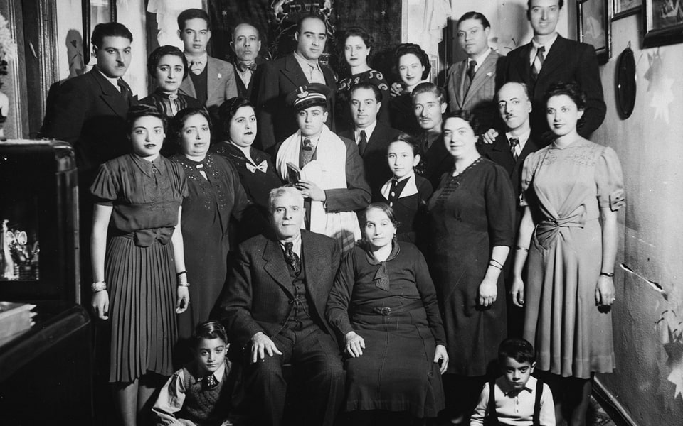 A Greek Jewish family gathers for a group portrait surrounding a bar mitzvah boy clad in a tallit. 1939