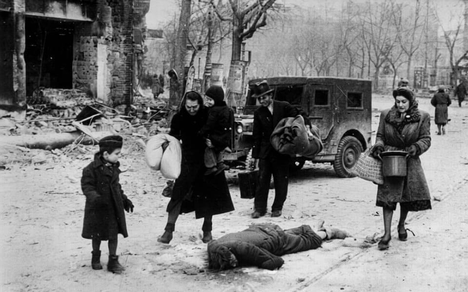 Civilians carry their personal belongings and walk past a dead person in the ghetto of Budapest in 1945